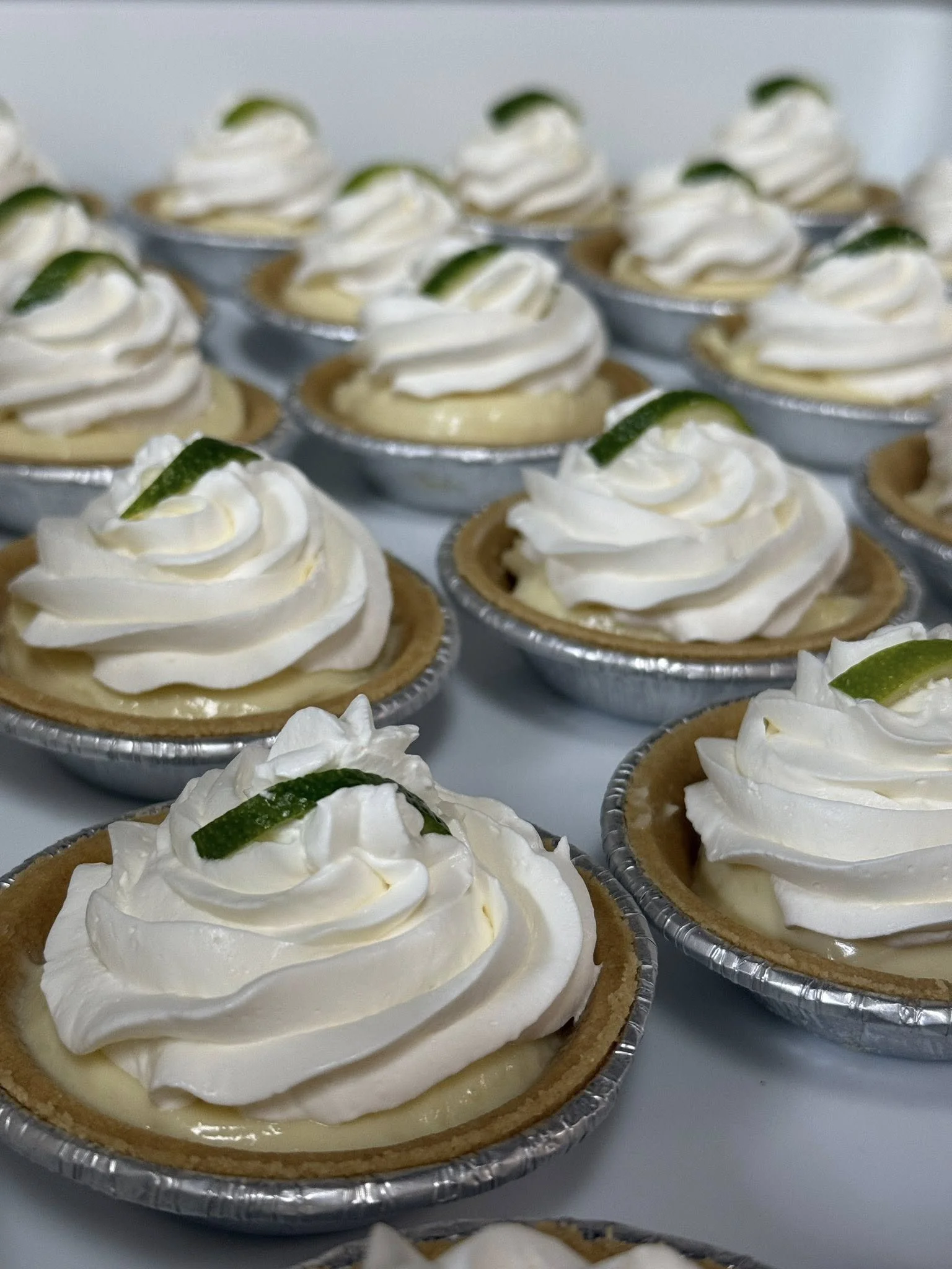 Lemon tartlets topped with whipped cream and lime zest garnishes, arranged in aluminum tart molds.