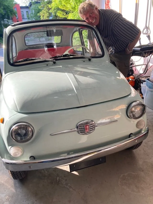 Man with gray hair and striped shirt smiling and leaning over a light green vintage Fiat 500 inside a garage.