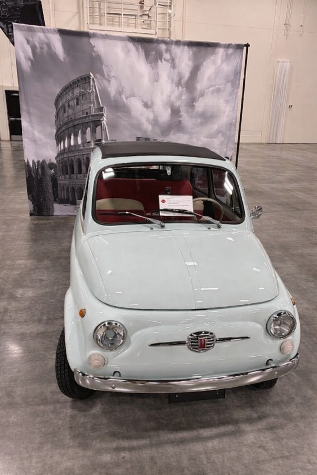 A vintage white Fiat 500 parked indoors with a red interior, in front of a black and white backdrop featuring the Colosseum and a cloudy sky.