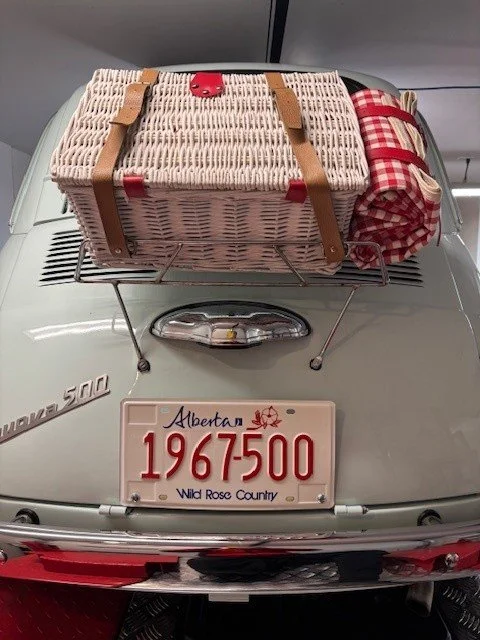 Vintage beige and red wicker picnic basket secured on the hood of a classic silver Volkswagen Beetle with an Alberta license plate.