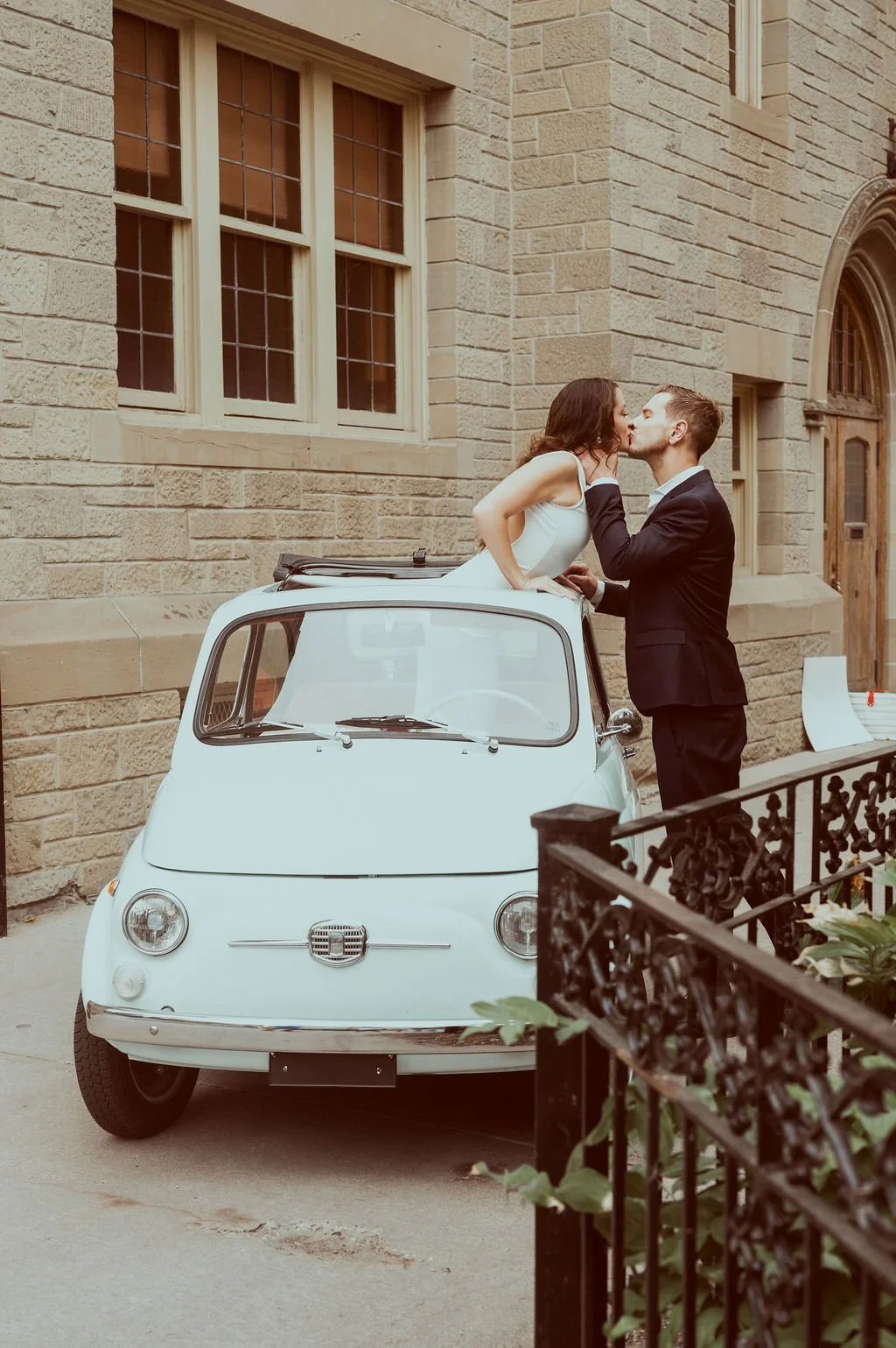 A couple sharing a kiss on top of a vintage white Fiat 500 parked outside a brick building with large windows.