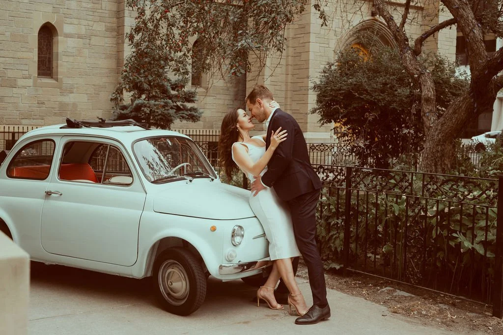 A couple dressed in formal wear embracing beside a vintage white Fiat 500 car in front of a church with gothic architecture.