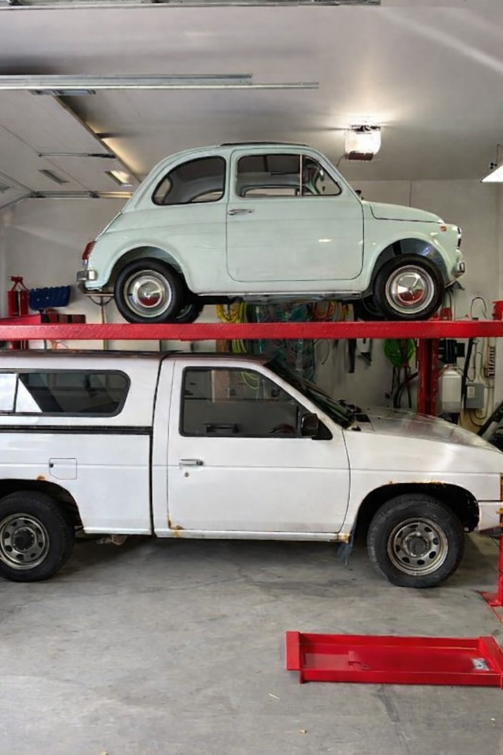 A vintage pale green car is parked on a red metal lift inside a garage, above a white vehicle with signs of rust and wear.