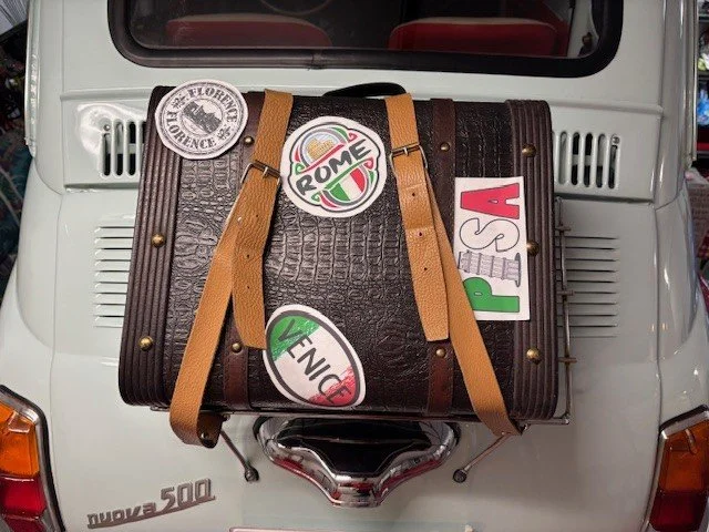 A white Vespa scooter with a leather suitcase on the front, decorated with colorful stickers including one with the word 'Rome', a 'Venice' sticker, and other travel-related stickers.