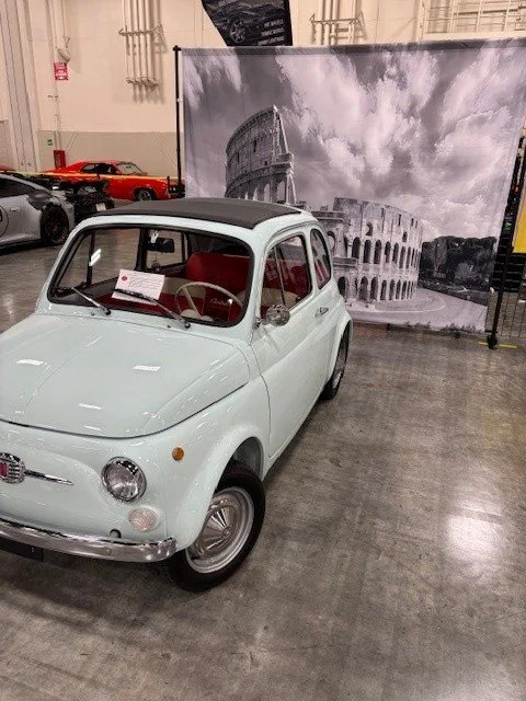 A vintage light blue Fiat 500 with a black roof on display indoors, with a large black-and-white photo of the Roman Colosseum in the background.