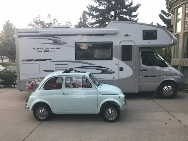 A vintage white Fiat 500 car parked in front of a large gray Vista Cruiser recreational vehicle on a paved driveway with trees and a house in the background.