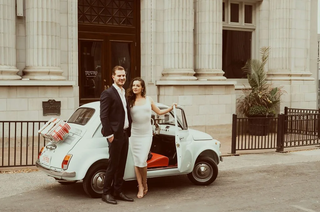 A man and woman in formal attire standing next to a vintage white Fiat 500 parked on a city street, in front of a building with large columns, with a gift box on the back of the car.