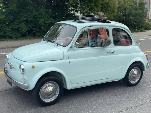 A person waving from the driver’s seat of a small, vintage, light blue Fiat 500 on a city street.