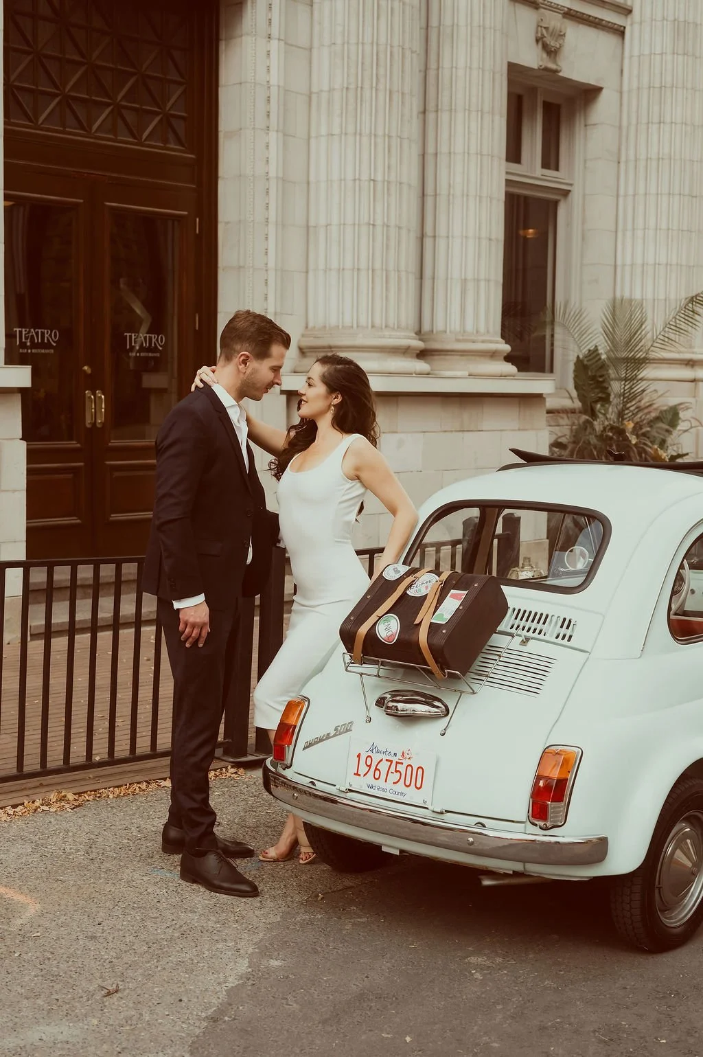 A man and woman dressed in formal attire standing close together near a vintage white Fiat 500 with luggage on its back, in front of a building with large columns and wooden doors.