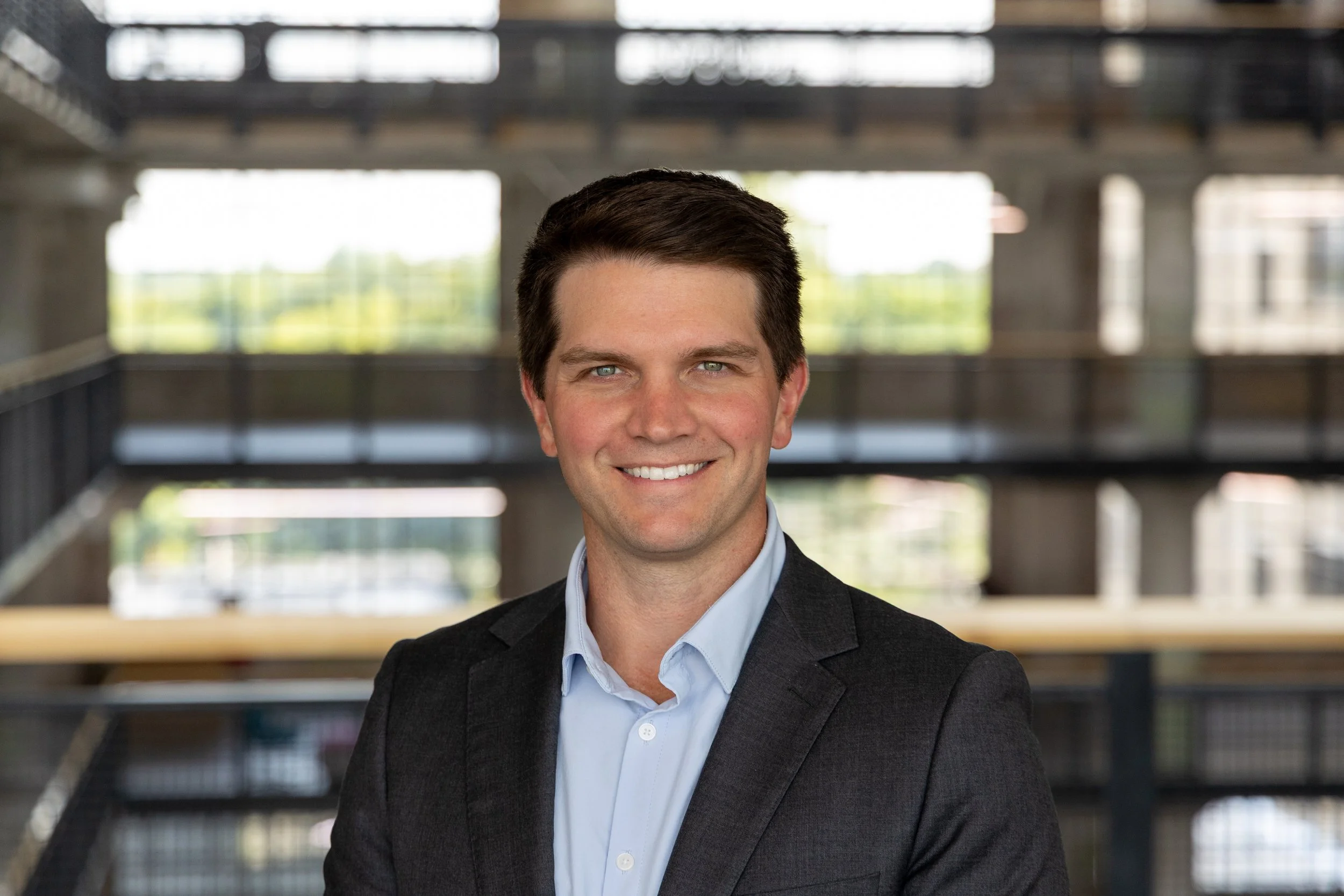 A man with dark brown hair and a suit smiling in an office building with an industrial interior and multiple levels.