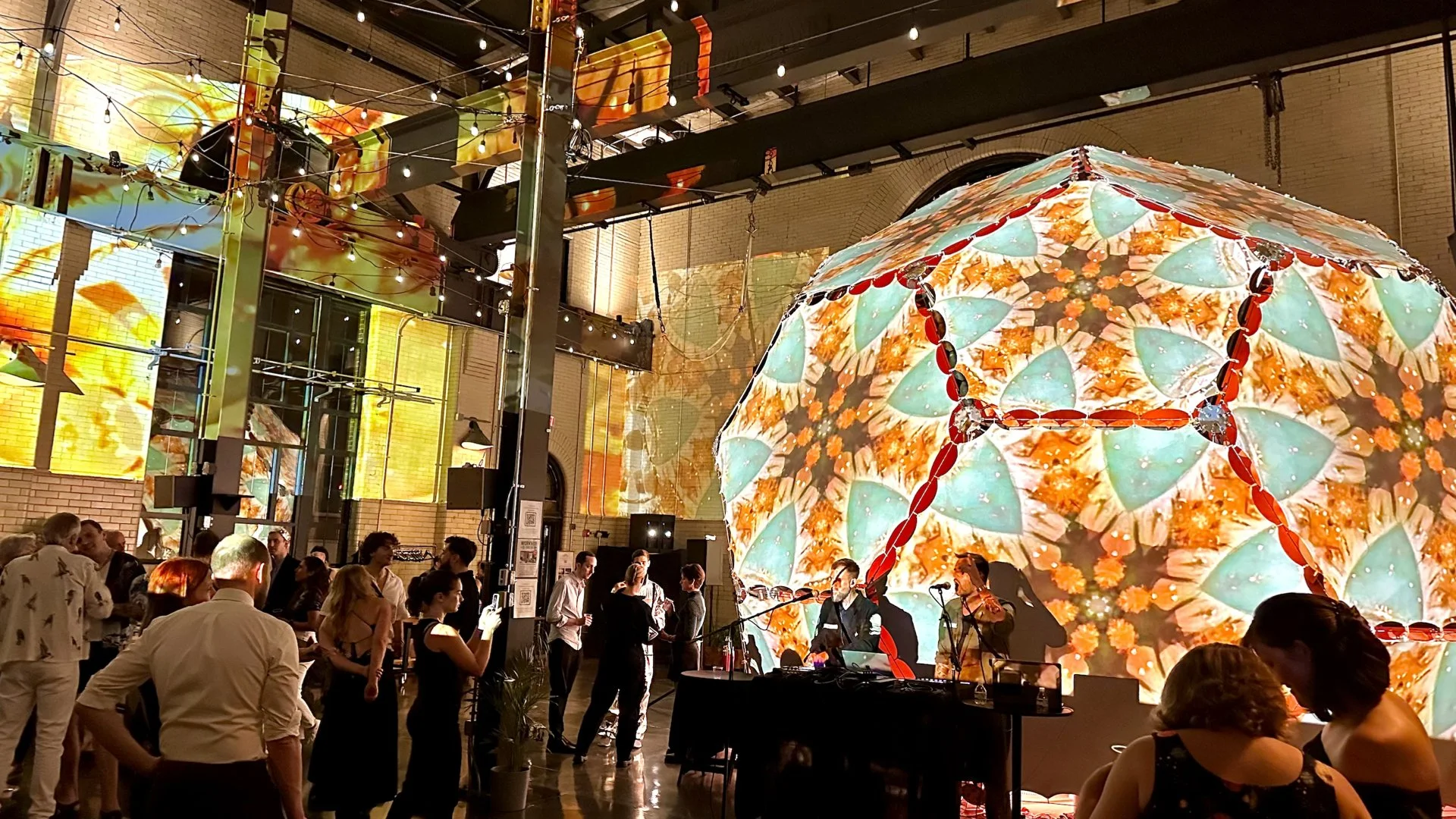 An indoor event with a large illuminated art installation resembling a giant kaleidoscope or umbrella with colorful floral patterns, and smaller screens displaying abstract visuals. People are socializing, some taking photos, and a DJ setup is visibl