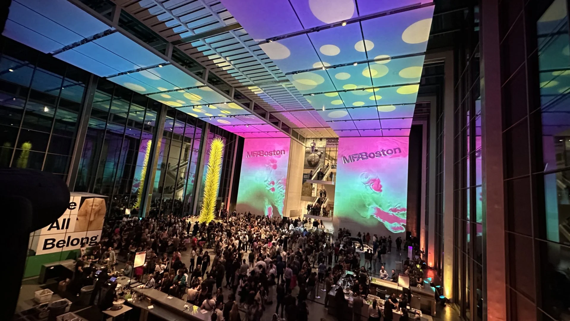 A large, modern indoor event space filled with a crowd of people. There are tall glass walls and a ceiling with colorful, illuminated patterns. Two large screens display the text 'MFA Boston,' and there is a tall yellow illuminated sculpture in the c