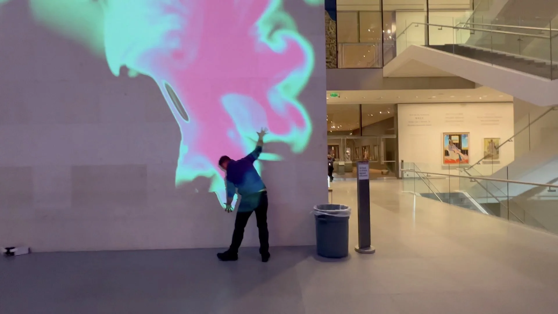 A man standing in a museum or gallery, reaching out towards a large colorful digital art display on the wall with swirling pink, blue, and green patterns.