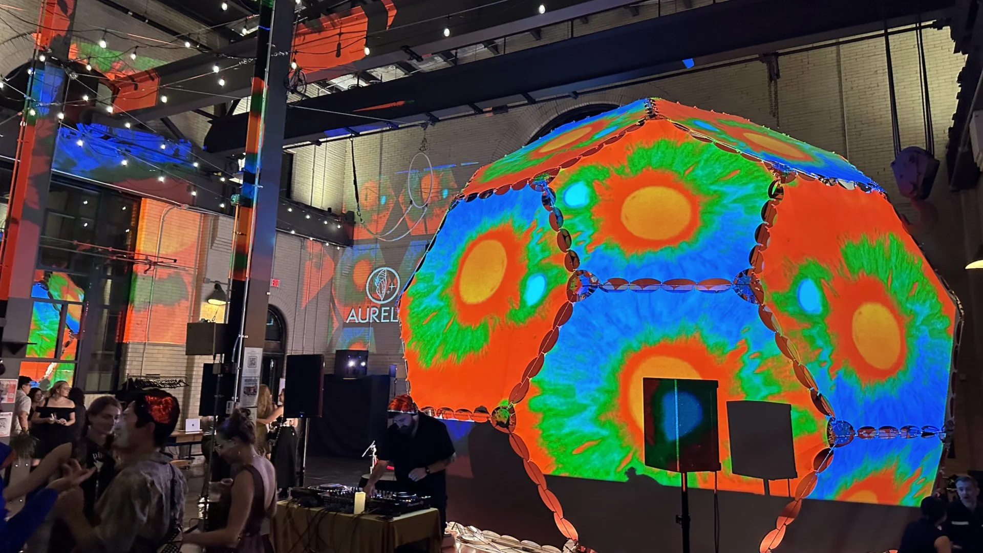 An indoor art installation featuring a large, illuminated geodesic dome with colorful, psychedelic patterns projected onto its surface, surrounded by a crowd of people.