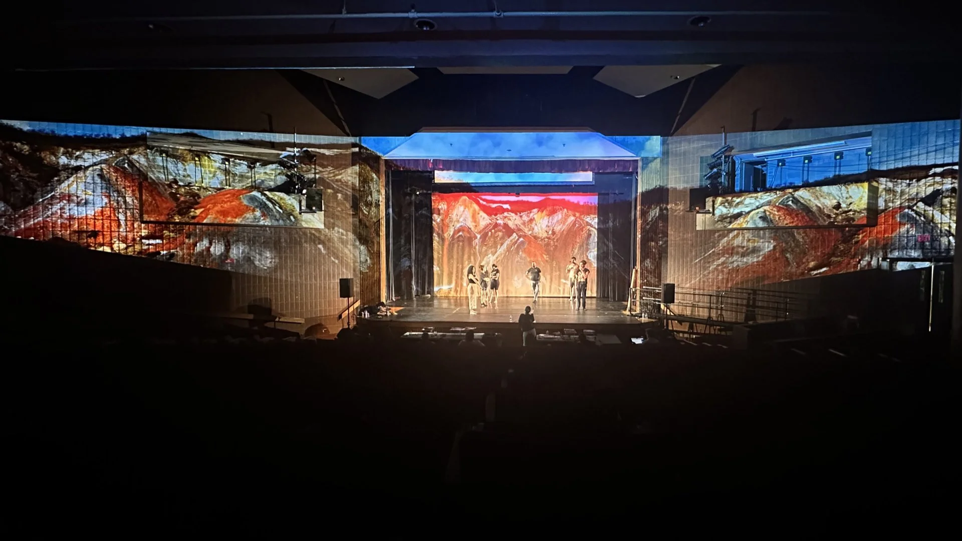 Empty theater stage with large colorful mountain landscape backdrop, surrounded by dark seating area, with five people standing on stage.