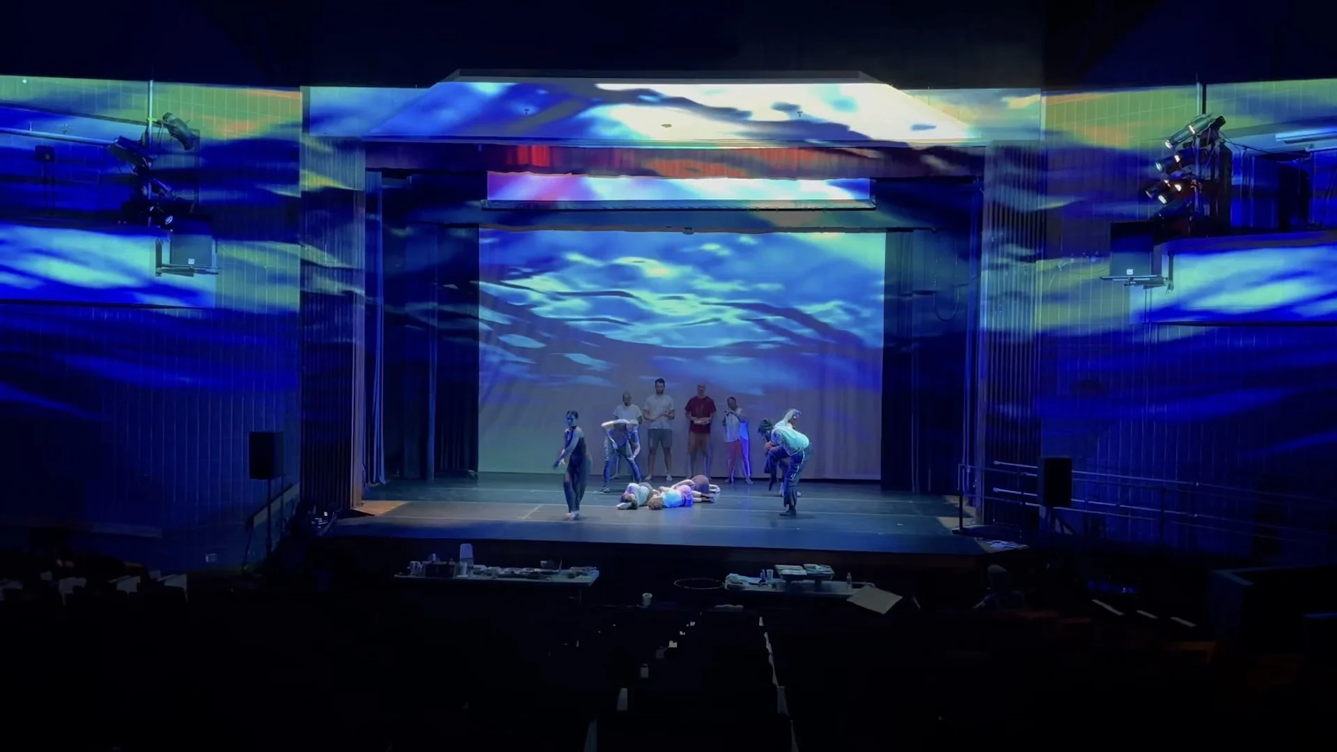 Stage with several performers dancing and lying on the floor, with blue ocean-like visuals projected on the backdrop and sidewalls.