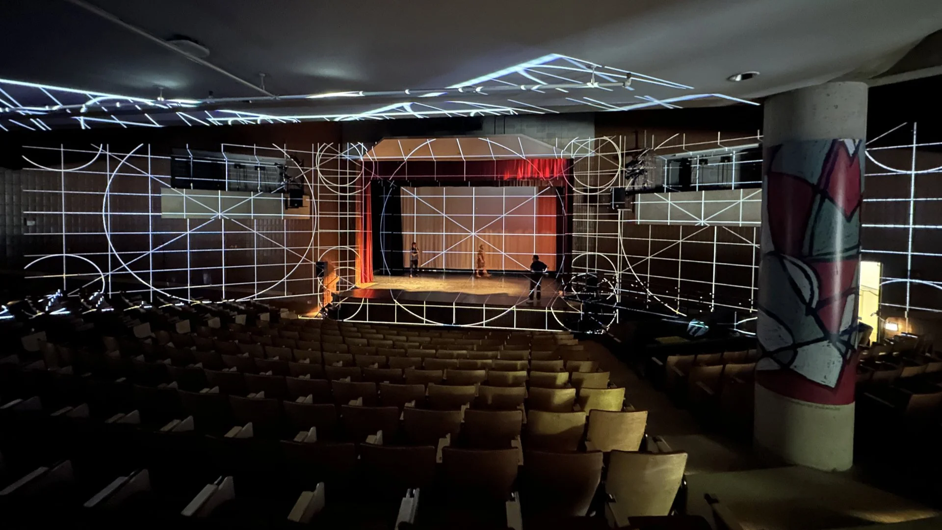 Empty theater with chairs facing a stage, illuminated by white lines forming geometric patterns on the walls, ceiling, and stage. People are setting up or rehearsing on stage, with some equipment visible.