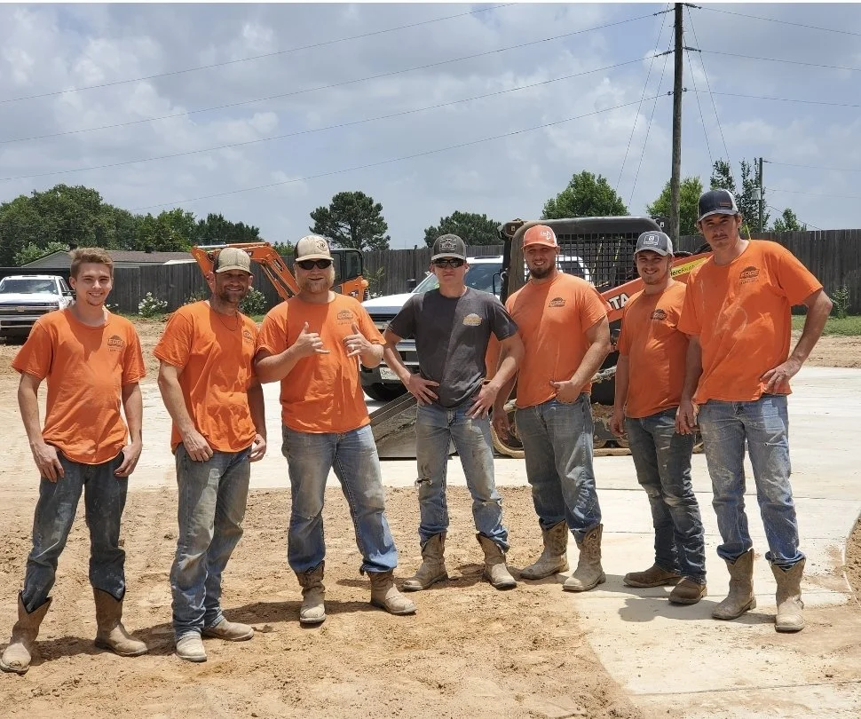 Group of seven construction workers standing on a construction site, posing for a photo with construction equipment and vehicles in the background, some making hand gestures.
