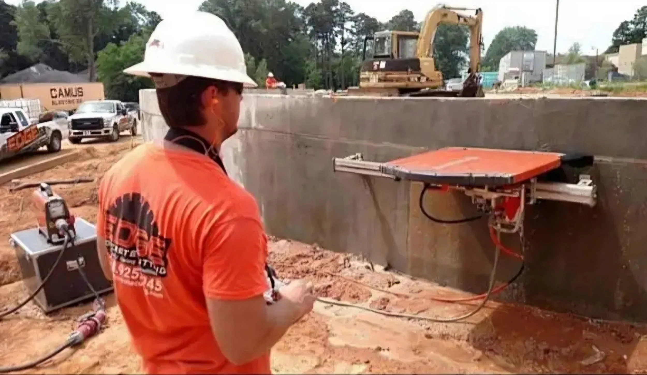 Construction worker wearing an orange shirt and white hard hat operating equipment at a construction site with an excavator, trucks, and construction materials in the background.