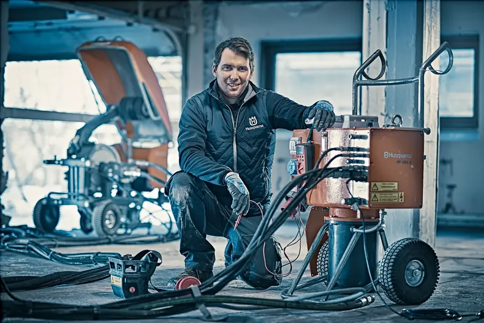 A young man using welding equipment inside a construction site. He is kneeling, smiling, and wearing work gloves and a jacket with the Husqvarna logo. Construction tools and machinery are visible in the background.
