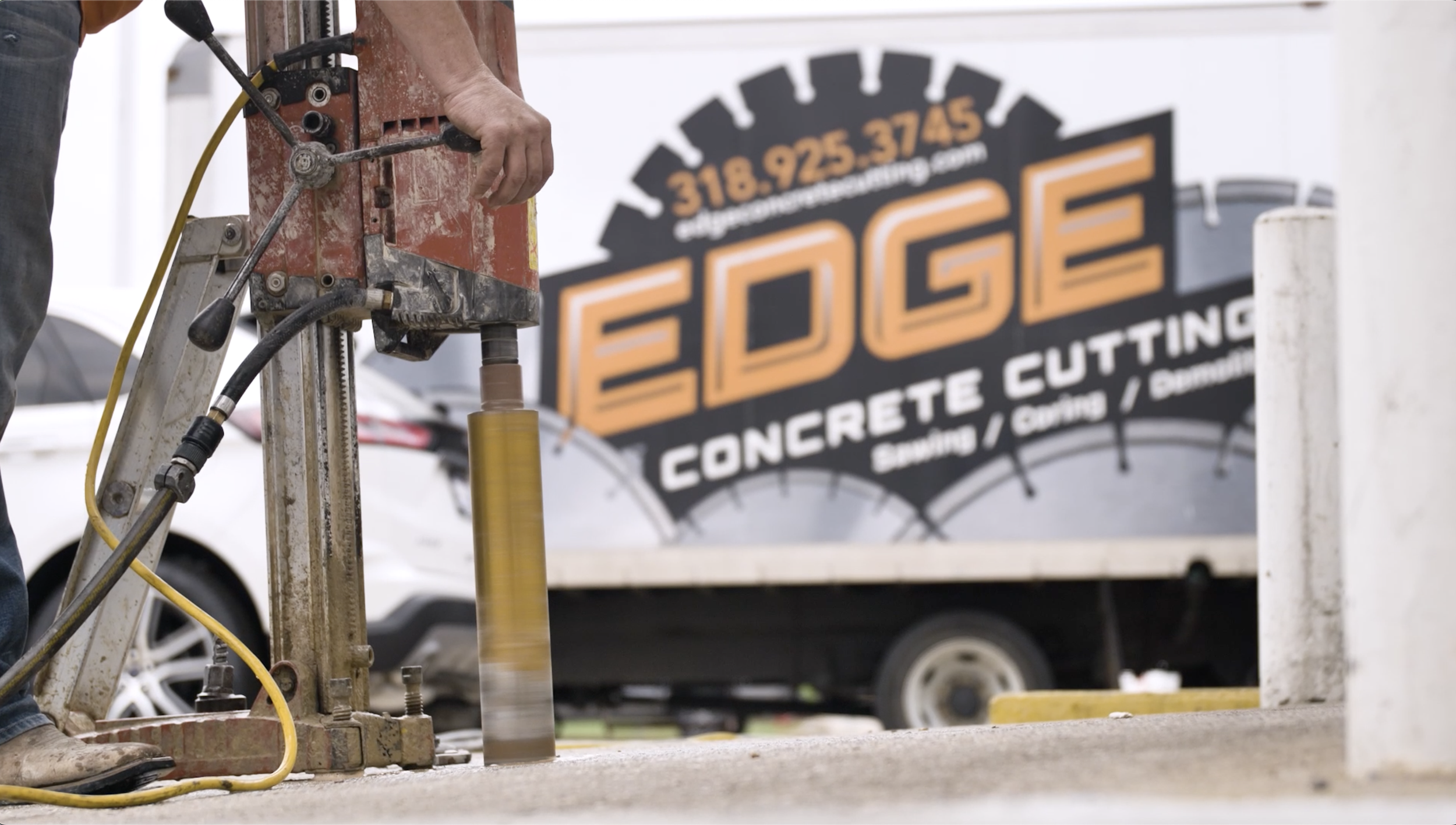 A construction worker operating a core drill on a concrete surface with a background sign for Edge Concrete Cutting.