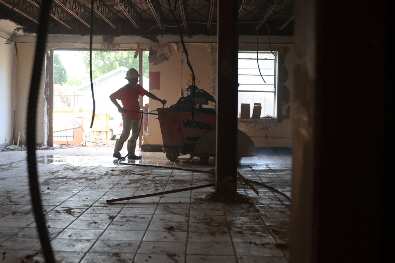 A construction worker operating a large floor grinder inside a partially demolished room with exposed walls and a window, with construction materials and debris on the floor.