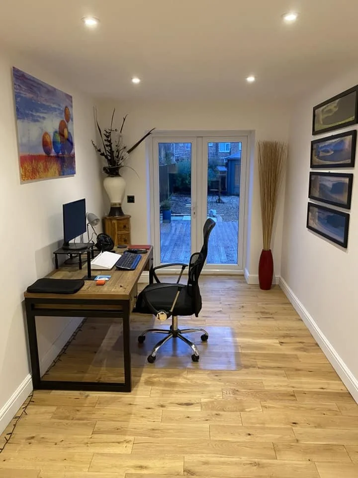 A home office with a wooden desk, a computer monitor, a black office chair, and a closed laptop. There is a large vase with decorative branches, a small wooden side table, and a large red vase with tall sticks. The room has hardwood floors, a set of