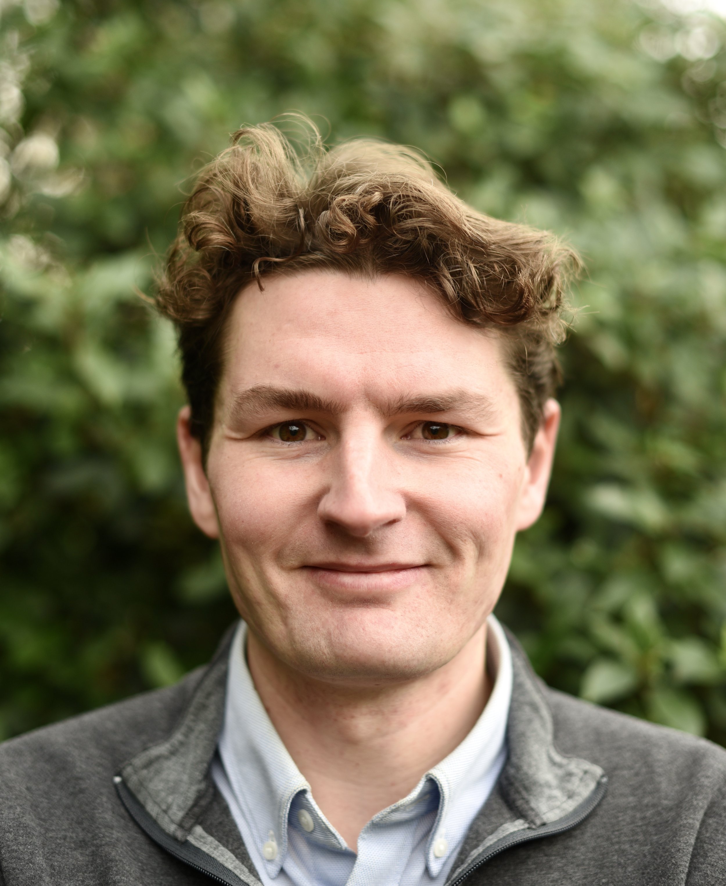 Close-up of a smiling young man with curly brown hair, wearing a gray jacket and light blue collared shirt, with a blurred green leafy background.