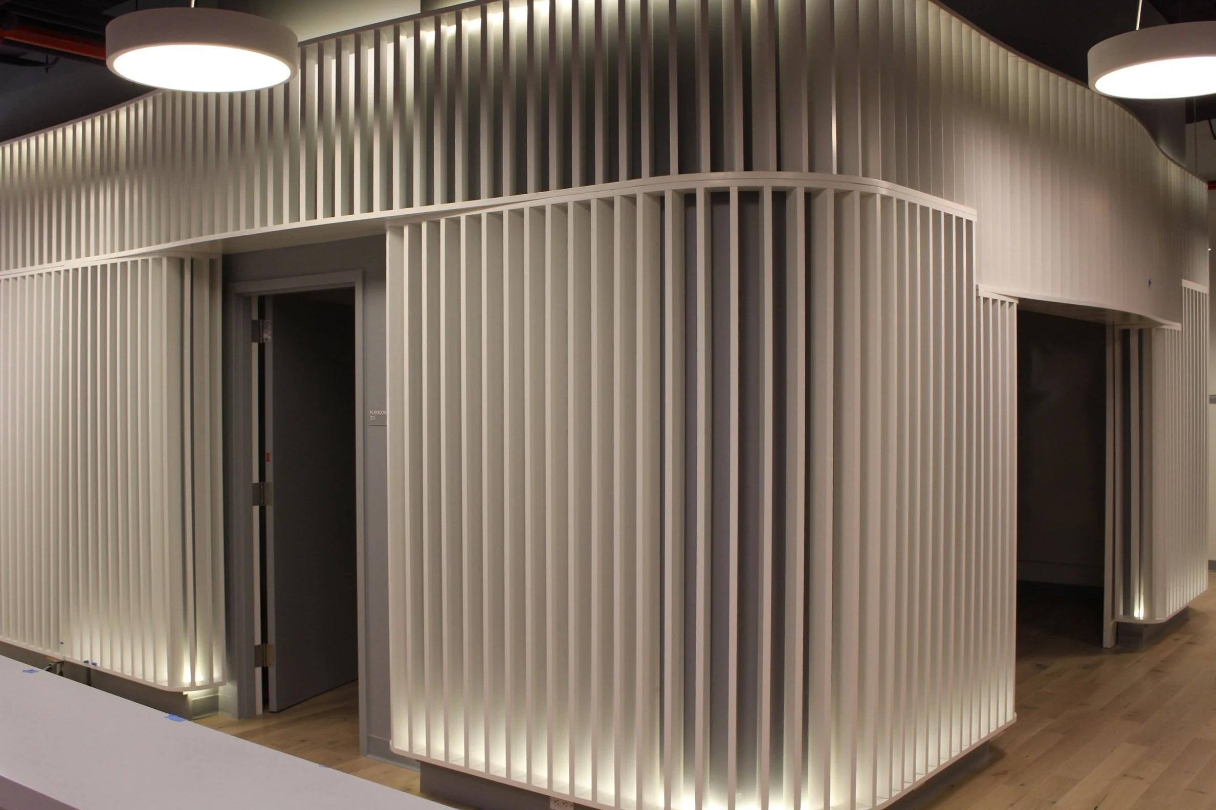 Interior view of education project the Kennedy School, with curved white walls featuring vertical slats and two partially visible doors, illuminated by overhead lighting and floor lighting.