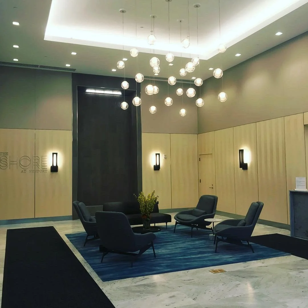 Residential project lobby with a seating area featuring four gray armchairs arranged around a coffee table on a blue rug. The walls are wood-paneled with black sconces and a large glass chandelier.