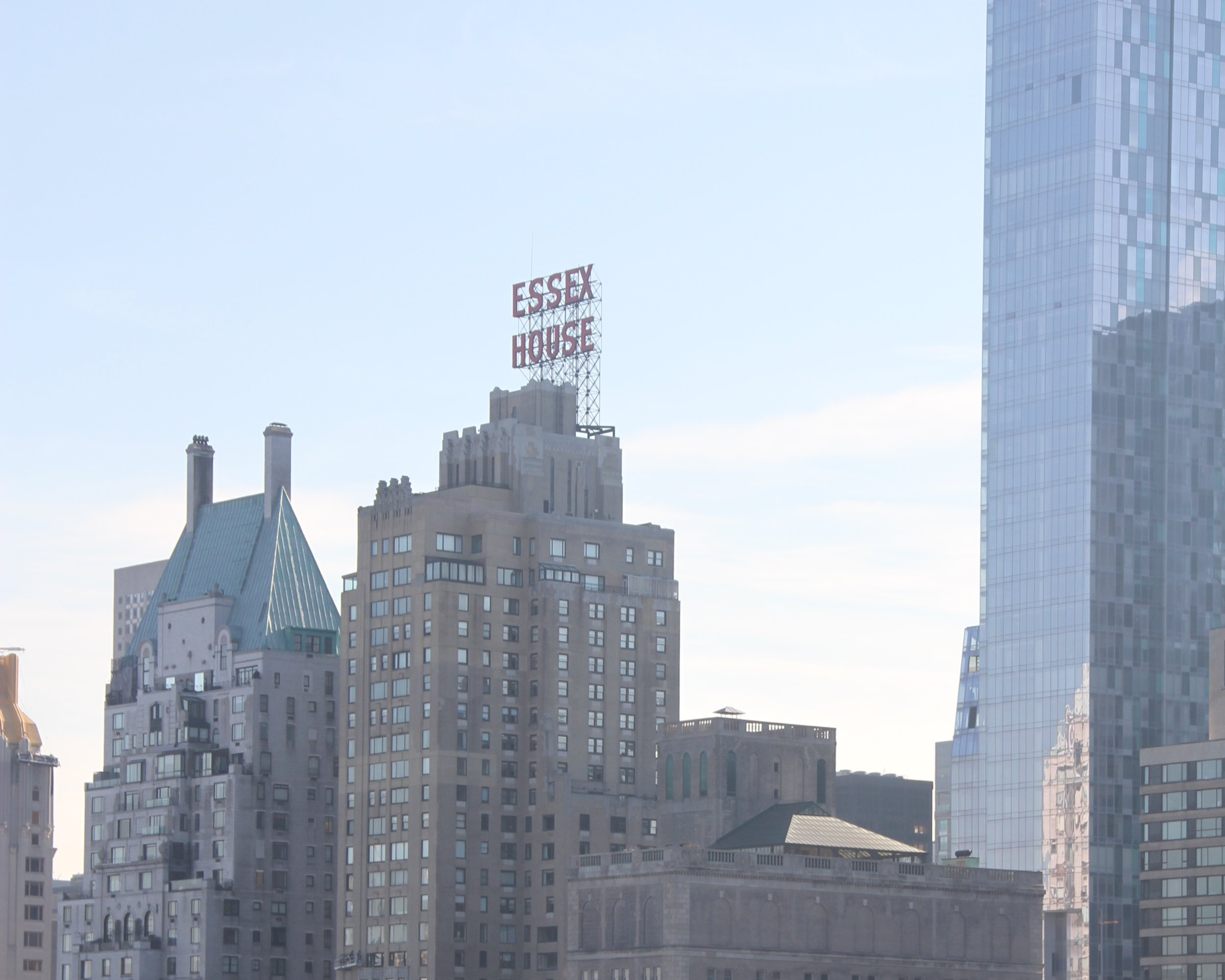 City skyline featuring the exterior of the Essex House building, a hospitality project. The skyscraper is topped with a red Essex House sign under a clear blue sky.
