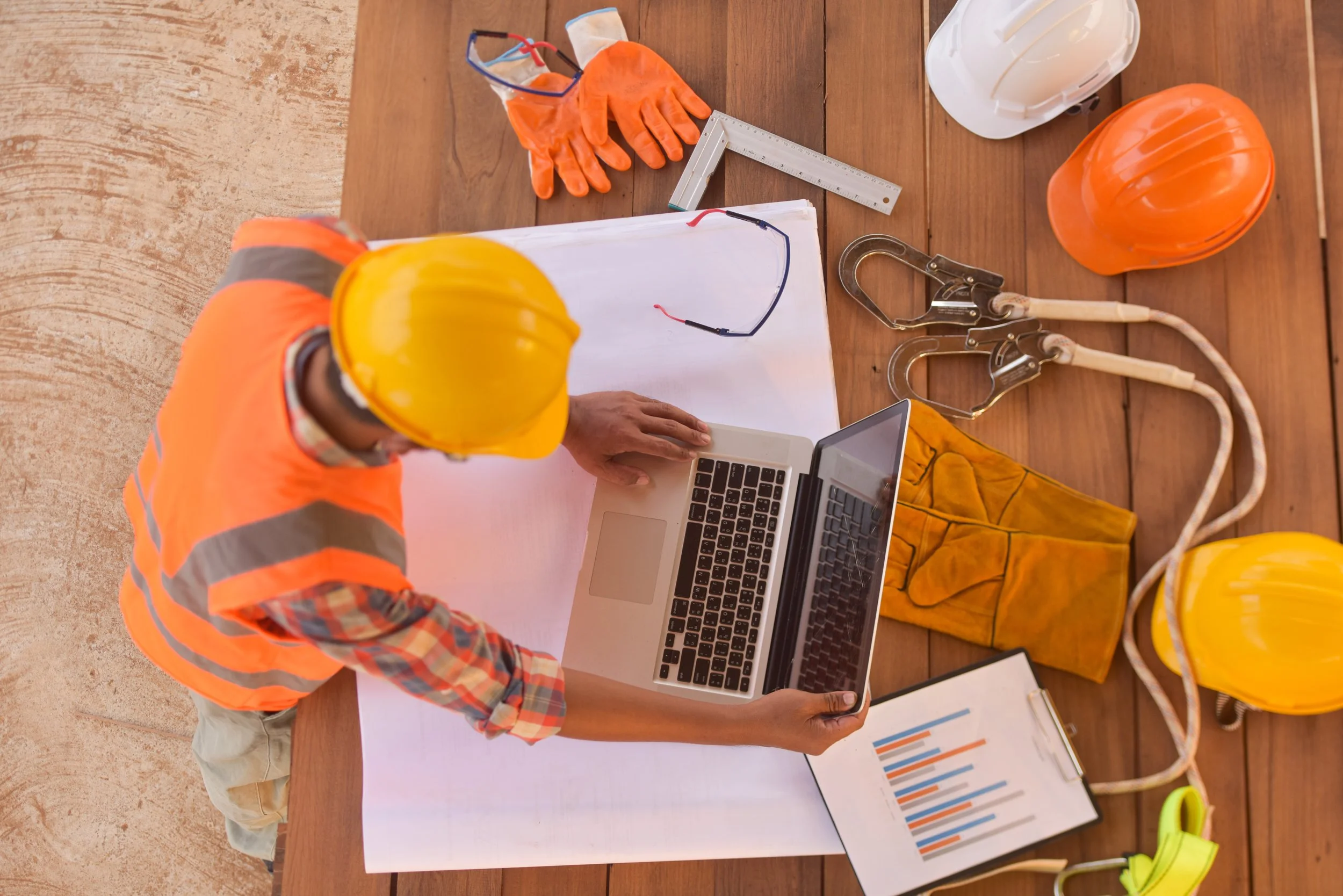 A construction worker in a safety helmet and orange safety vest using a laptop at a wooden table with construction tools, helmet, safety gloves, and chart documents.