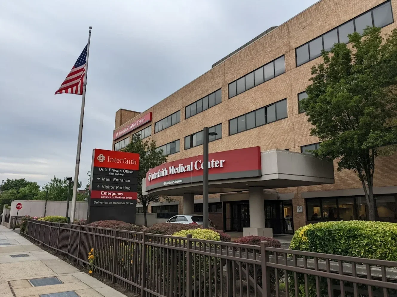 Exterior view of Interfaith Medical Center, a brown brick multi-story building with a red sign, main entrance under a concrete canopy, American flag flying, trees, and a sidewalk with a metal fence and shrubs.