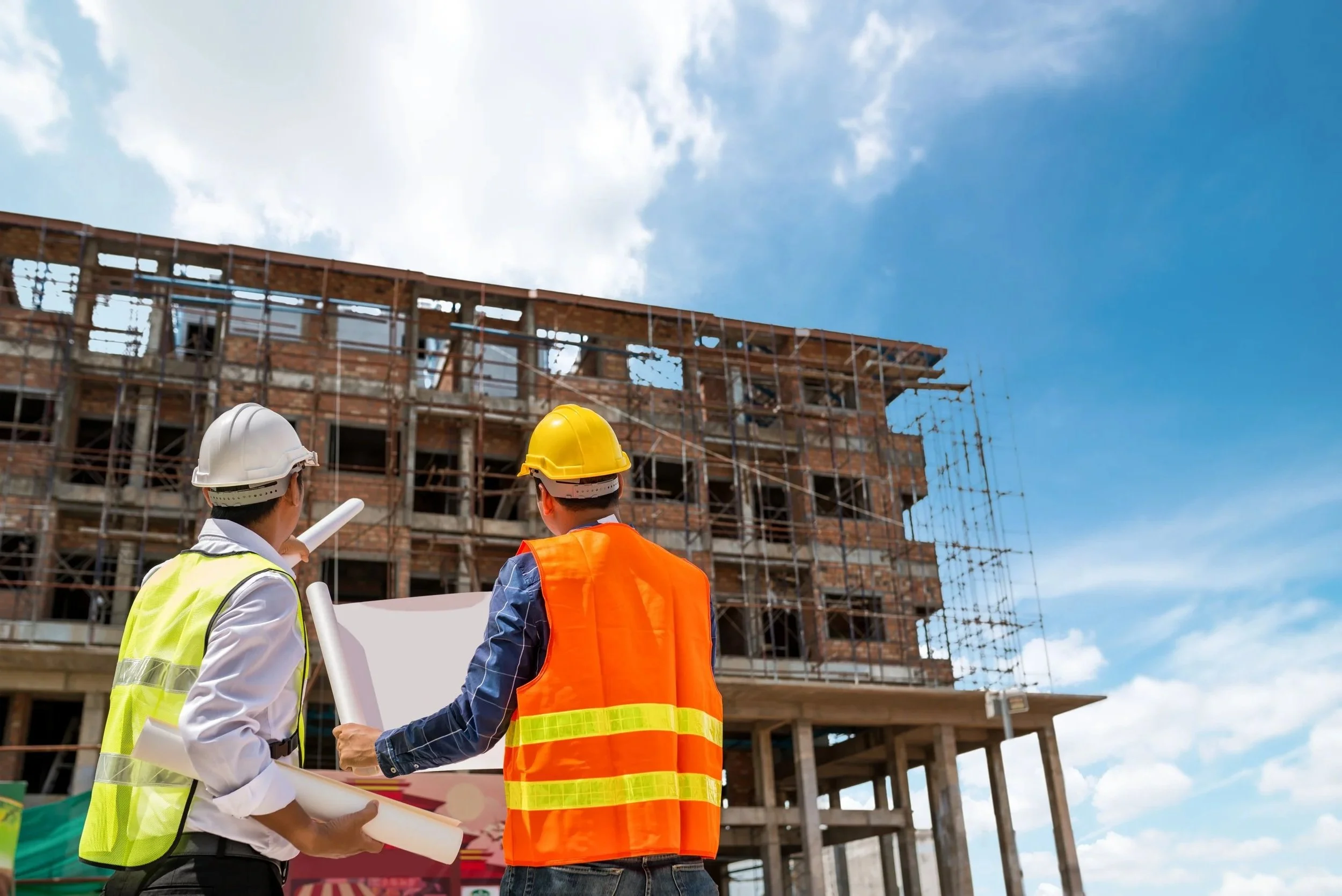 Link to “evaluation & advisory” page. Two construction workers in vests and hard hats talking while looking at a building with scaffolding.