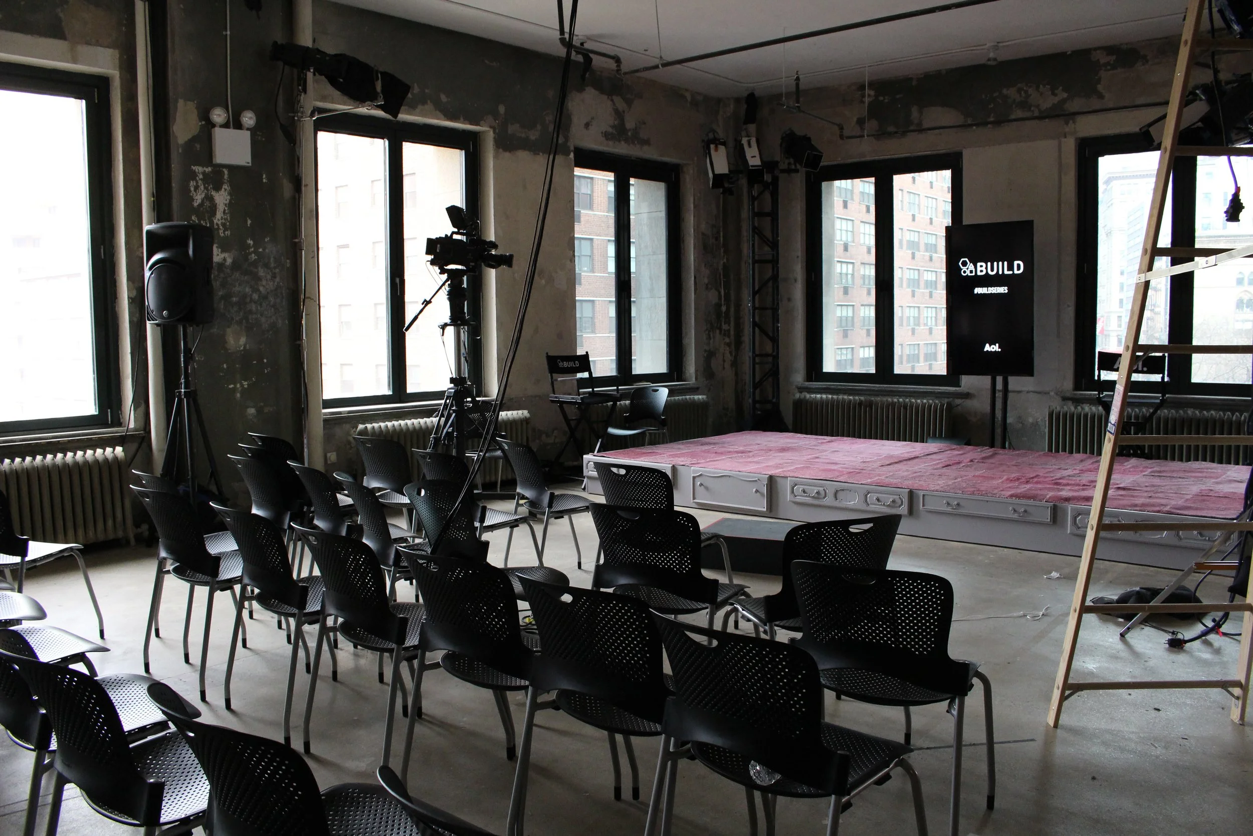 Empty event space at commercial project AOL Build, with chairs arranged in rows, a small stage with a pink cover, camera equipment, and a large screen displaying 'BUILD' in an urban loft with large windows.