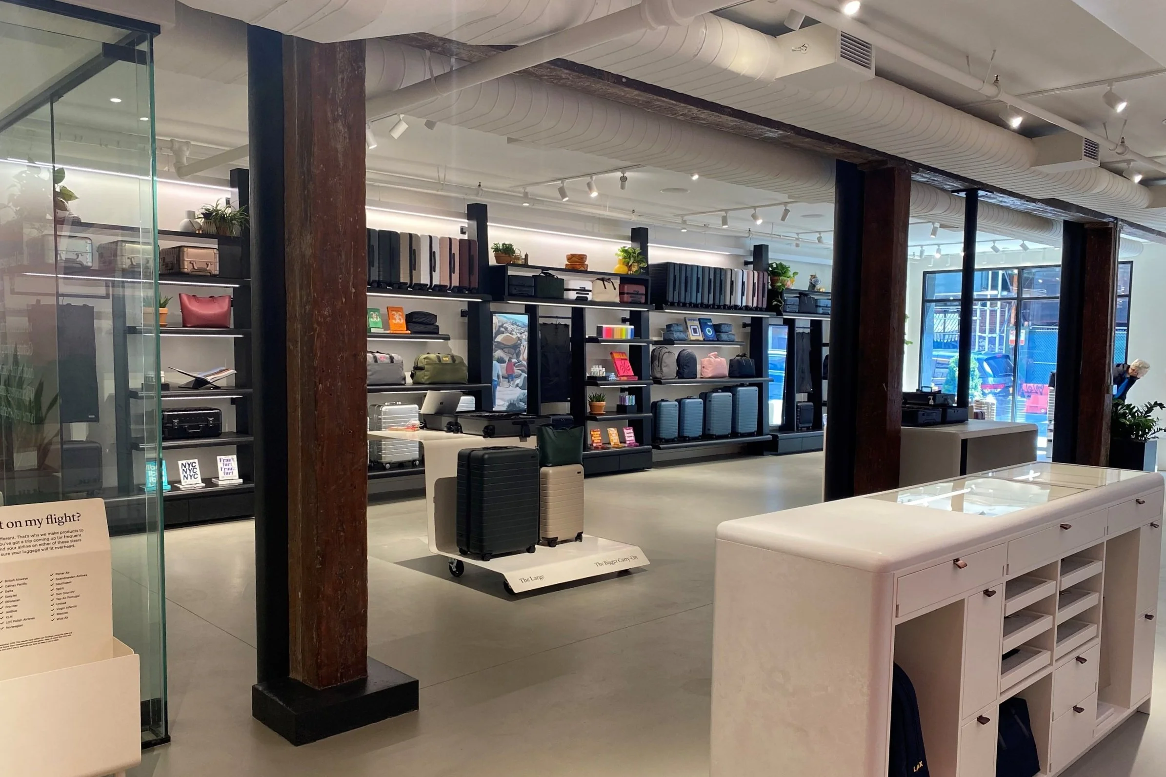 Interior of retail store Away Travel displaying luggage, handbags, and accessories on black shelves. The store has large glass windows, white ceiling with exposed ductwork, and wooden support columns.