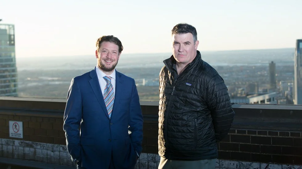 Quest Builders Group executives Eric Derector, Principle, and Patrick Fitzpatrick, Principle, standing on a rooftop with a cityscape in the background, one in a blue suit and the other in a black jacket.