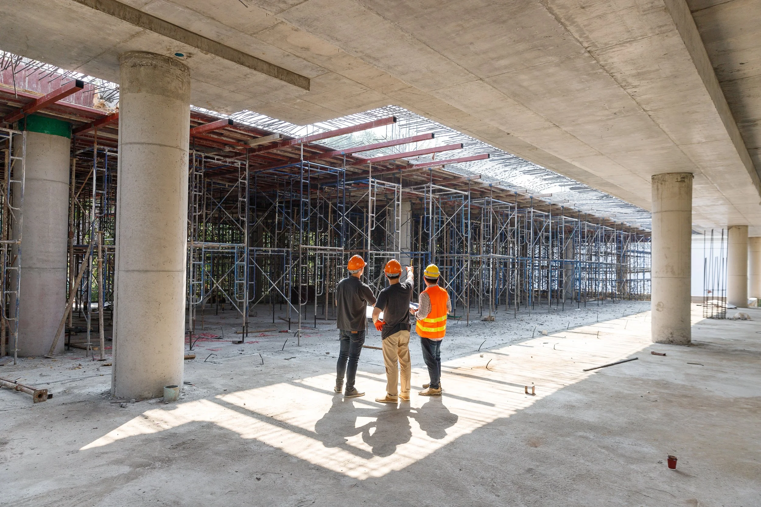 Three construction workers wearing orange and yellow safety helmets are standing and discussing a building under construction. The building has scaffolding and concrete columns, with a large open space.