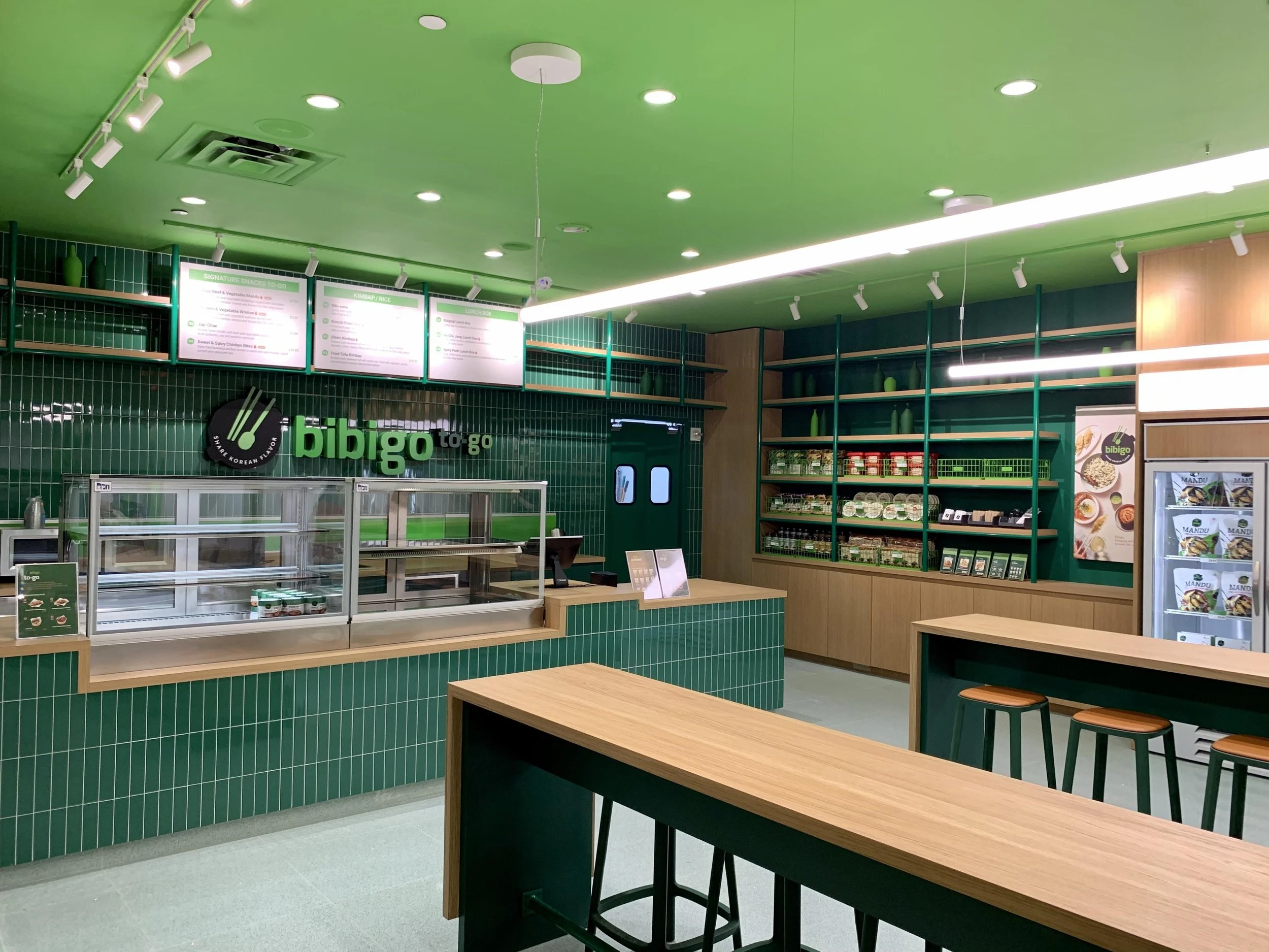 Interior of restaurant project, Bibigo, with green tiled walls, a counter with glass display cases, a wooden table, stools, shelves with products, and a refrigerated display with packaged food.