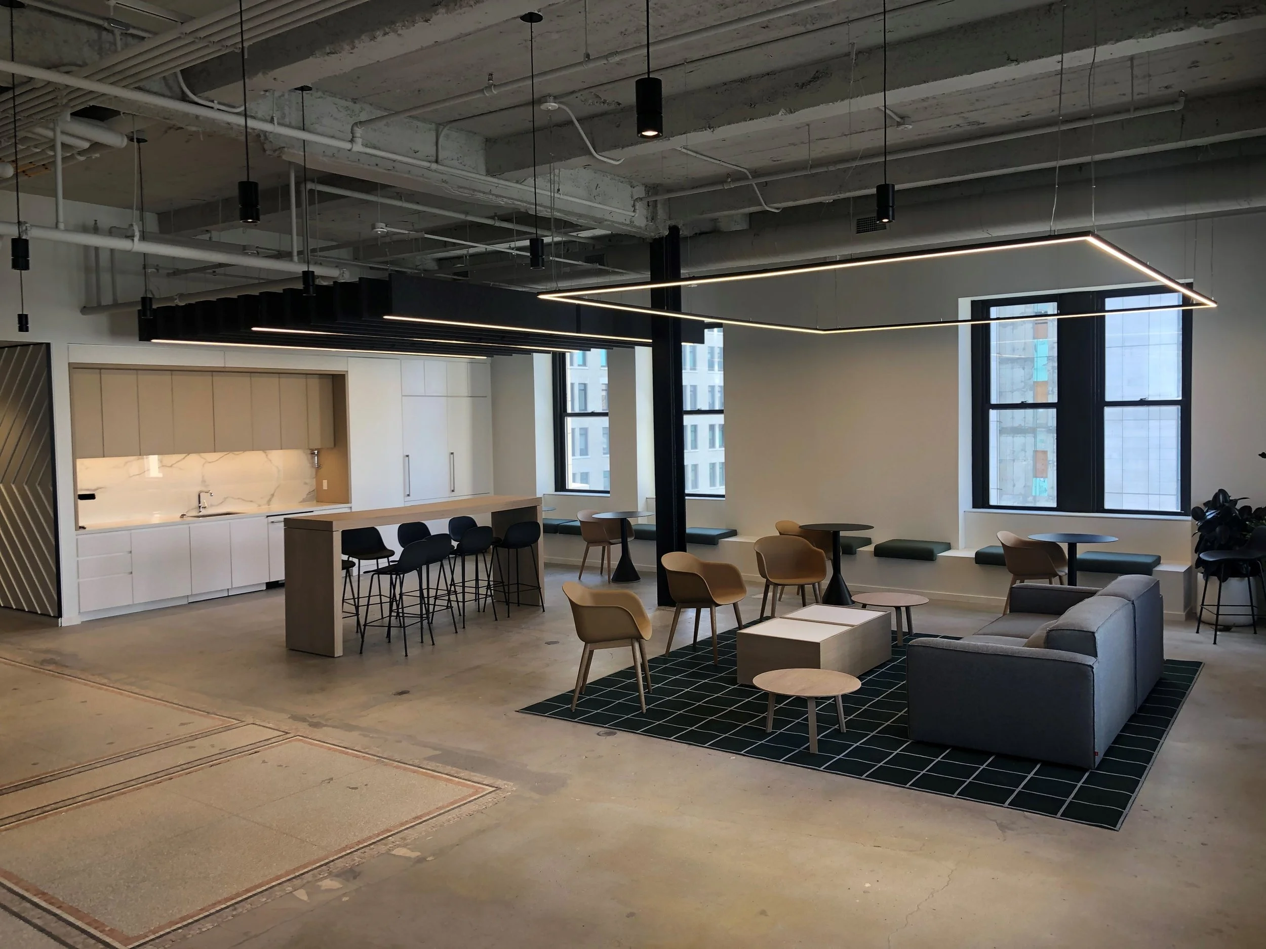 Interior view of landlord project Cove Properties. Modern office lounge area with a gray sofa, beige chairs, coffee tables, and a kitchenette with white cabinets and a marble backsplash, contemporary lighting fixtures and large windows.