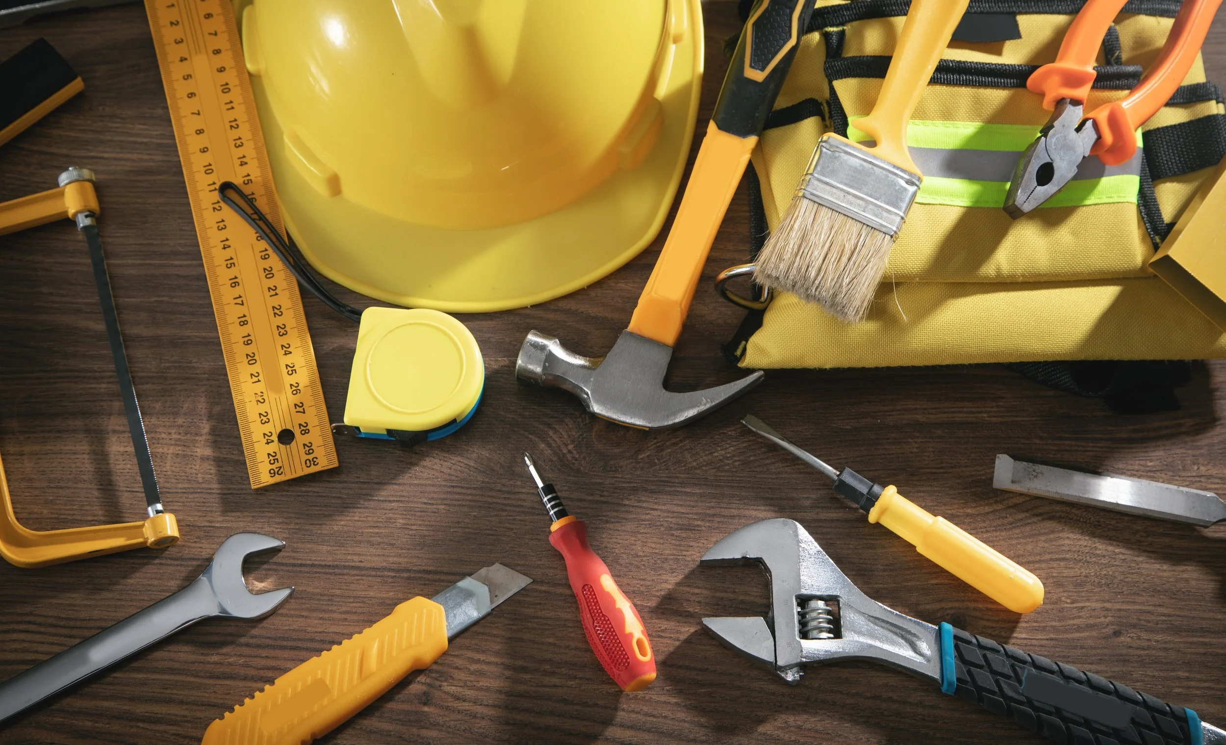 Link to "general contracting" page. Various construction tools and safety gear, including a yellow hard hat, measuring tape, hammer, screwdrivers, wrenches, pliers, a yellow tool bag, and a yellow safety vest on a wooden surface.