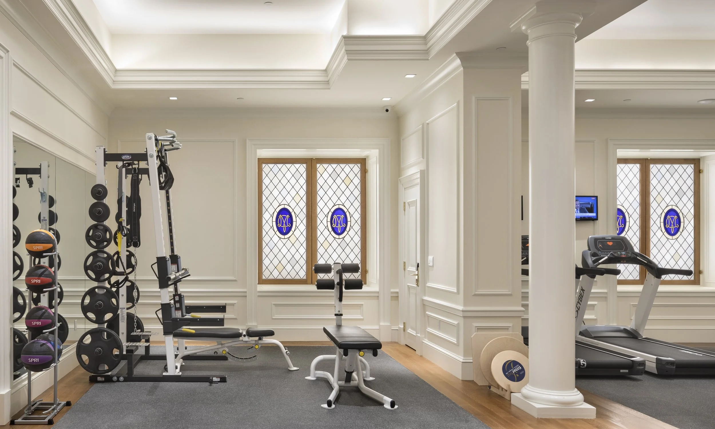 Interior view of the gym at hospitality project the Metropolitan Club, with workout equipment including dumbbells, a weight rack, an adjustable bench, an exercise bike, and a treadmill in a room with large stained glass windows depicting their logo.