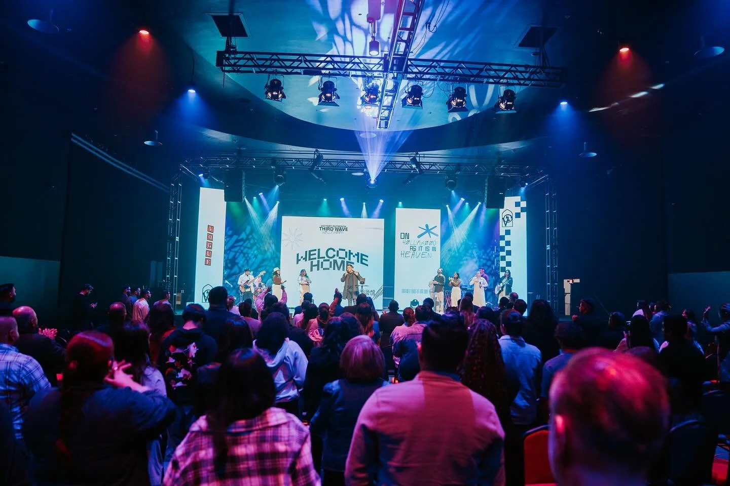 People attending a concert or worship service in a dimly lit venue with stage lights, some standing with hands raised, one man with glasses in the foreground raising his hand.