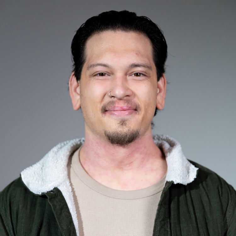 A man with dark hair, a goatee, and a nose piercing, wearing a gray shirt and a black jacket with a white lining, smiling at the camera against a plain gray background.