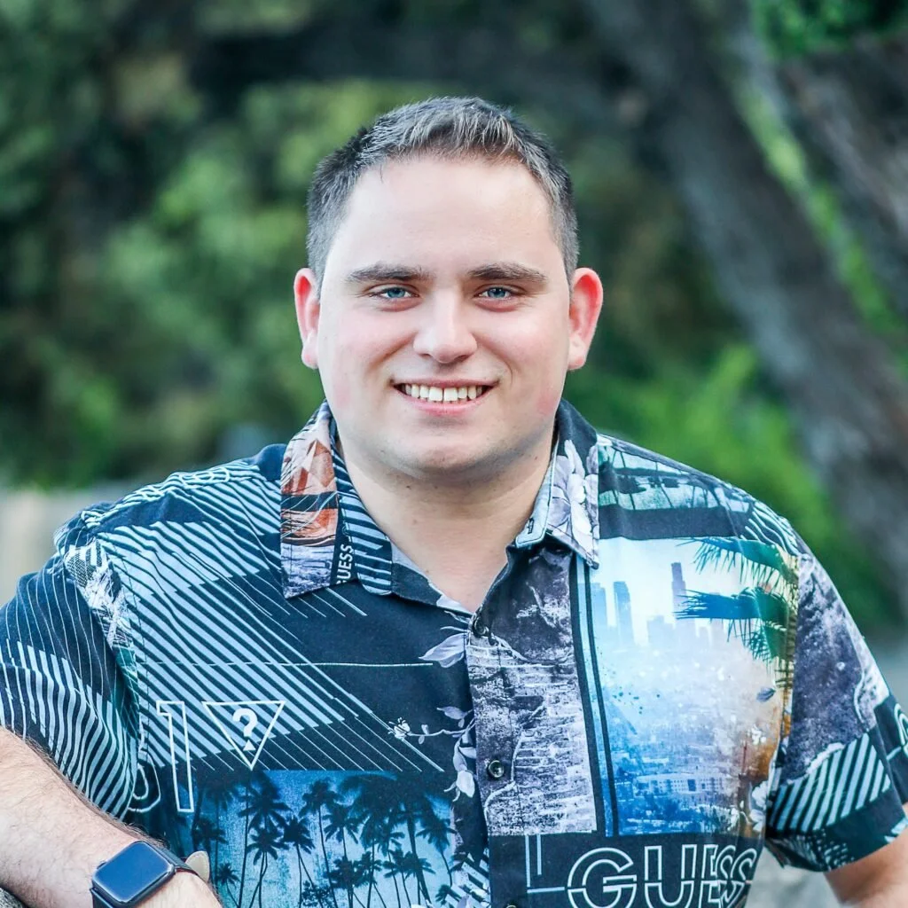A young man with short brown hair and blue eyes smiling outdoors in a green, wooded area, wearing a colorful Hawaiian shirt.