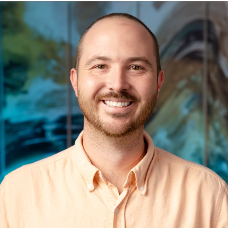 Smiling man with short hair and beard wearing a beige collared shirt, standing in front of a colorful abstract background.