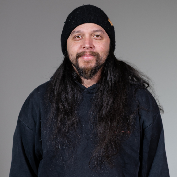 A man with long black hair, a beard, and mustache, wearing a black beanie and a black hoodie, smiling at the camera against a plain gray background.