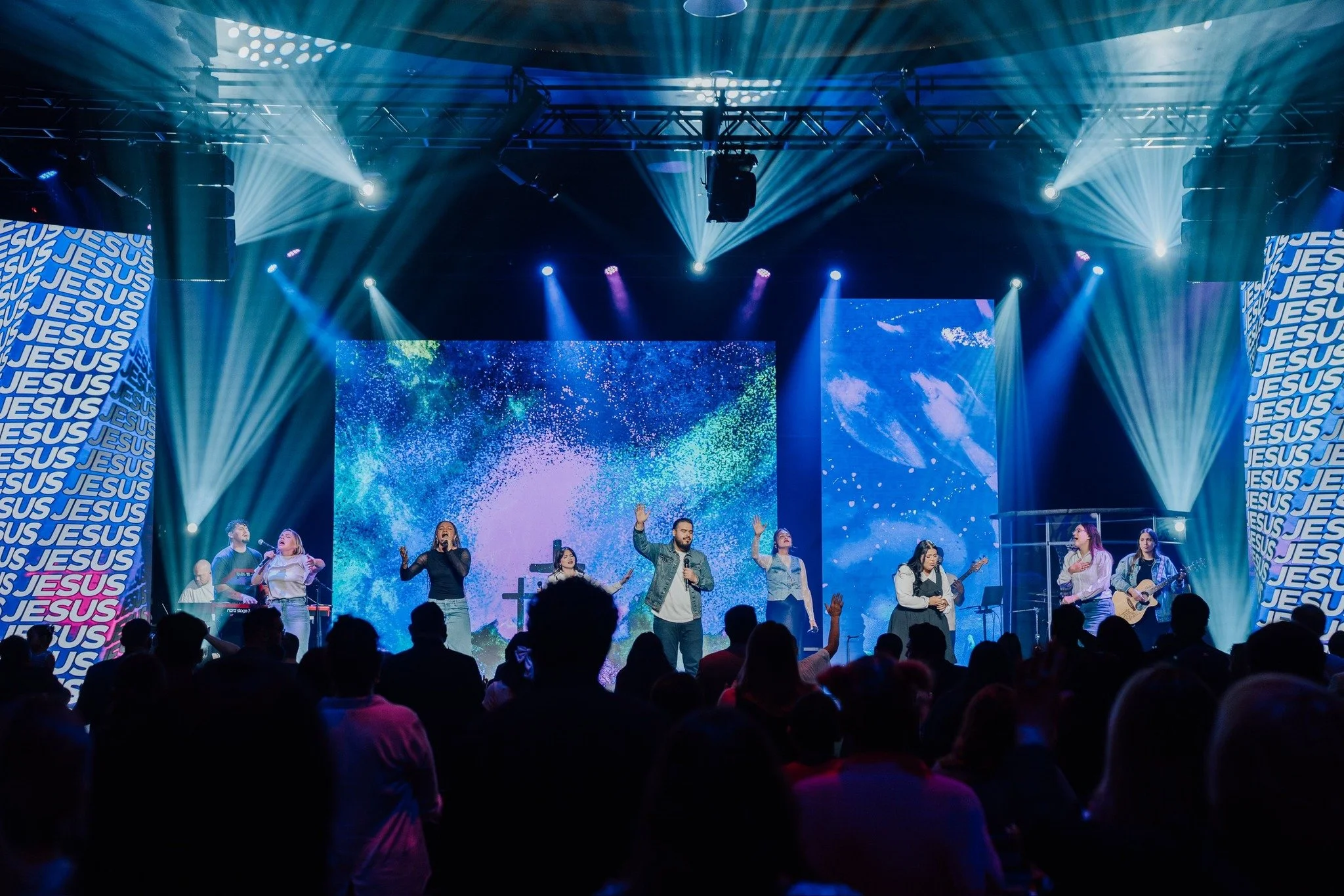 A group of musicians and singers performing on a stage with colorful lights and screens displaying abstract visuals, during a religious event with a crowd in the audience.