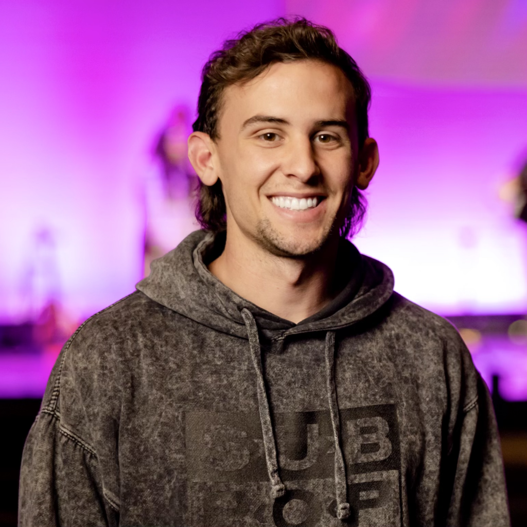A young man with light skin and shoulder-length wavy brown hair, smiling, wearing a gray hoodie, standing in front of a colorful, blurred background with purple and pink hues.