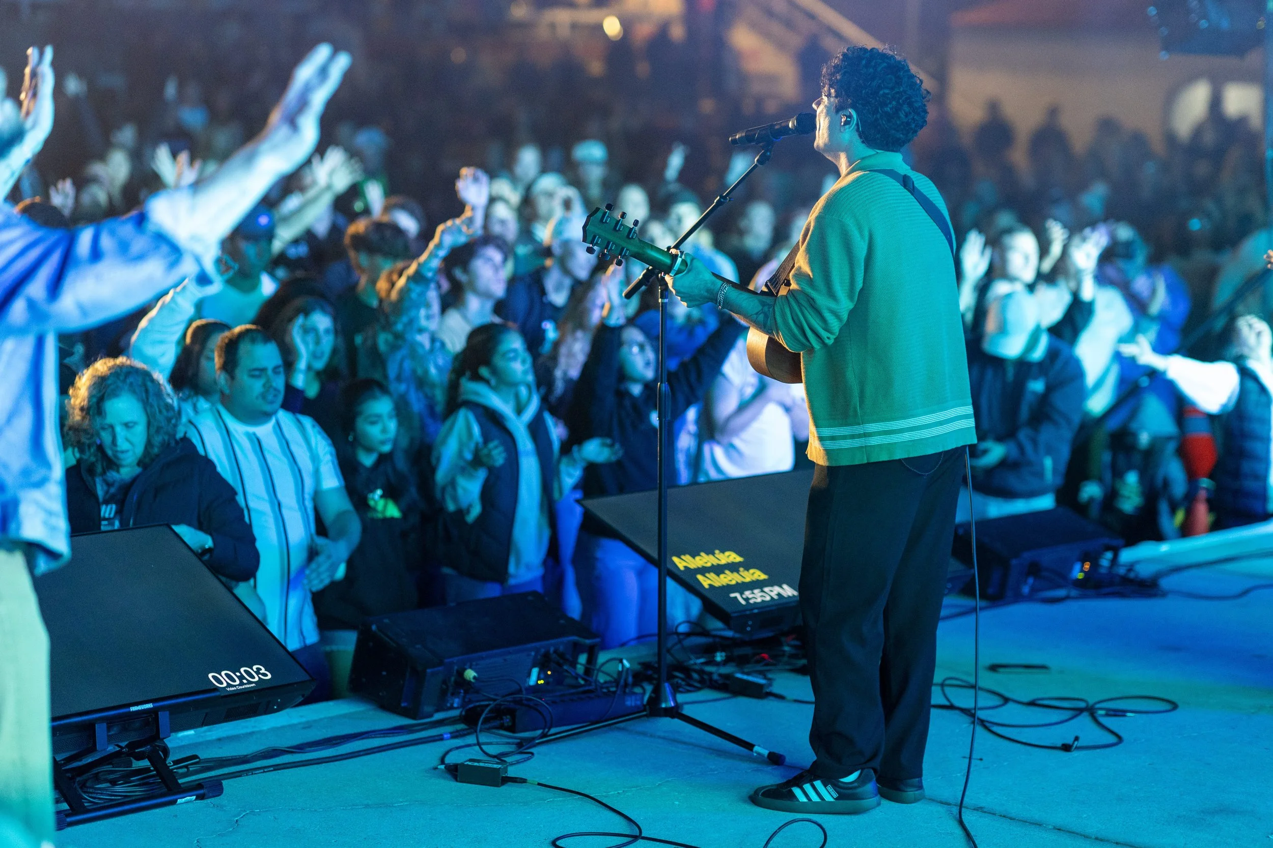 A musician wearing a green jacket and black Adidas slides performs with a guitar on stage in front of a large crowd at night. The audience has their hands raised and is engaged with the music.